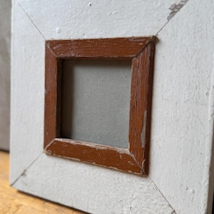 Op de afbeelding: Vierkante, wit geschilderde houten fotolijst met een kleiner, bruin gebeitst binnenframe. De lijst heeft een verweerde, vintage uitstraling met zichtbare slijtage. Het binnenframe is vierkant en het buitenframe is schuin.