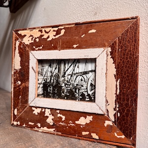 May include: A rectangular wooden picture frame with a distressed, aged appearance. The frame is primarily brown with areas of peeling paint revealing a lighter color underneath. The framed image is a black and white photograph.