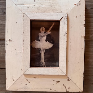 Könnte beinhalten: Ein rechteckiger Bilderrahmen aus abgenutztem, weiß gestrichenem Holz mit sichtbarer Maserung. Der Rahmen enthält ein Schwarz-Weiß-Foto einer Balletttänzerin in einem weißen Tutu, die in einer anmutigen Pose mit erhobenen Armen dargestellt ist.