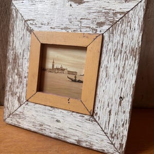 Op de afbeelding: Een vierkante fotolijst met een verweerde witte buitenrand en een gladde lichtbruine binnenrand. De lijst bevat een sepia-kleurige afdruk van een Venetiaans kanaal met een gondel, een persoon en gebouwen in de verte.