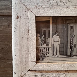 Può includere: Una cornice rettangolare con una finitura in vernice bianca usurata e una trama visibile delle venature del legno. La cornice contiene una fotografia in bianco e nero che ritrae diverse figure su un portico davanti a un edificio.
