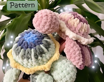 May include: Two crocheted turtle amigurumi toys. One is green, yellow, and blue, and the other is pink and white. The toys are held in a person's hand. The image includes the text "Pattern" and "@emmylaine_crochet".