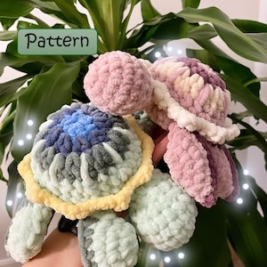 Könnte beinhalten: Zwei gehäkelte Amigurumi-Spielzeug-Schildkröten. Eine ist grün, gelb und blau, die andere ist rosa und weiß. Das Spielzeug wird in der Hand einer Person gehalten. Das Bild enthält den Text "Pattern" und "@emmylaine_crochet".