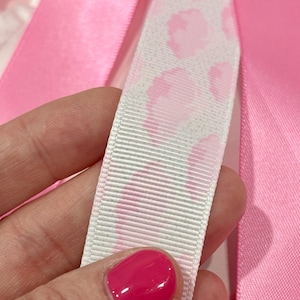 May include: A white grosgrain ribbon featuring a repeating pink leopard print pattern, positioned alongside smooth, solid pink satin ribbons. The textured white ribbon contrasts with the vibrant, glossy finish of the surrounding pink fabric strips.