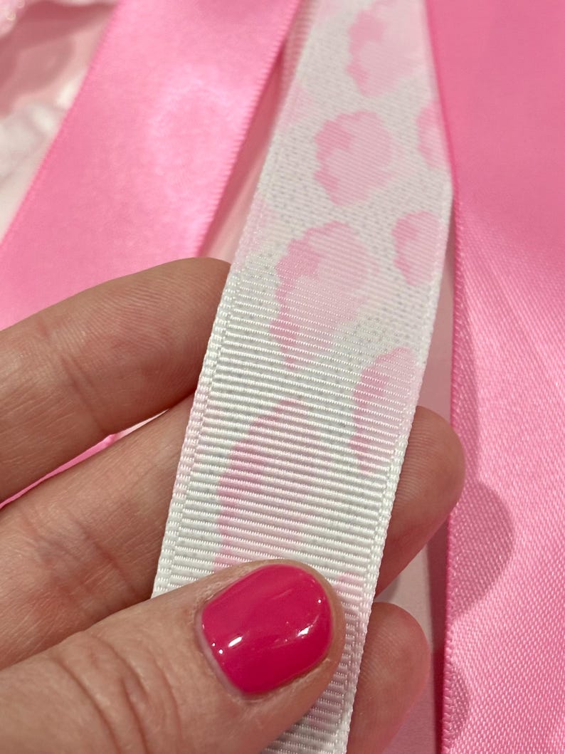 May include: A white grosgrain ribbon featuring a repeating pink leopard print pattern, positioned alongside smooth, solid pink satin ribbons. The textured white ribbon contrasts with the vibrant, glossy finish of the surrounding pink fabric strips.