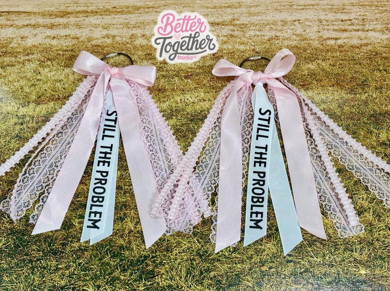 May include: Two pink hair accessories with bows and ribbons, one with a light blue ribbon that reads "STILL THE PROBLEM" in black text. The accessories have lace and are on a green and brown background.