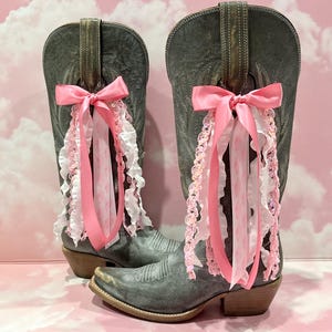 May include: A pair of distressed grey leather cowboy boots featuring decorative pink and white ribbon accents. Each boot is adorned with a large pink satin bow tied to the pull strap, with cascading ruffled ribbons and sequined trim hanging down.