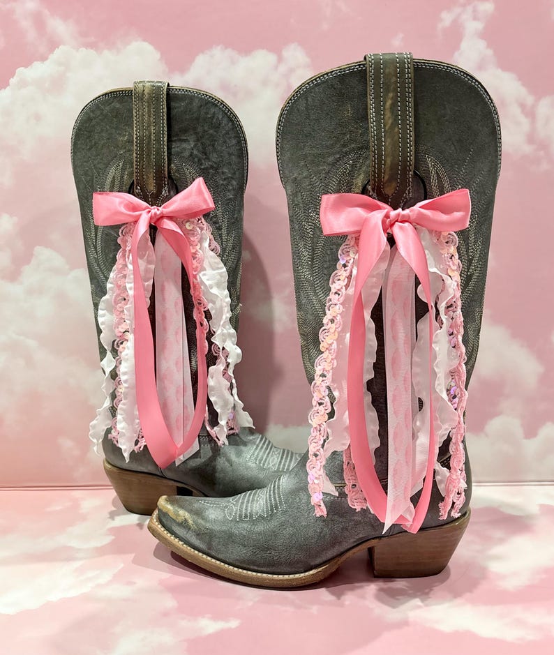 May include: A pair of distressed grey leather cowboy boots featuring decorative pink and white ribbon accents. Each boot is adorned with a large pink satin bow tied to the pull strap, with cascading ruffled ribbons and sequined trim hanging down.