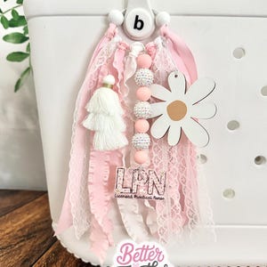 May include: A decorative bag charm featuring pink and white fabric ribbons, a white tassel, a string of pink and glittery beads, and a white daisy cutout. An acrylic charm displays the text LPN Licensed Practical Nurse in a floral pattern.