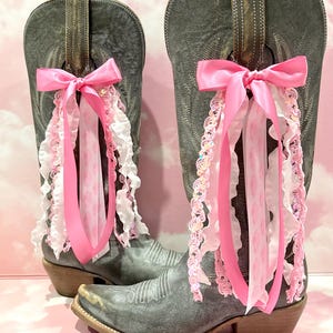 May include: A pair of distressed grey leather cowboy boots featuring decorative pink satin bows and cascading ribbons in shades of pink and white. The ribbons include ruffled fabric and sequined trim attached to the pull tabs of each boot.