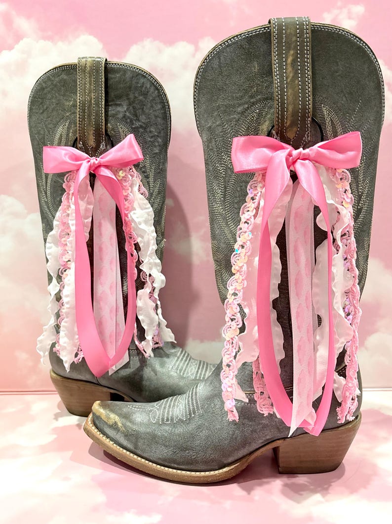 May include: A pair of distressed grey leather cowboy boots featuring decorative pink satin bows and cascading ribbons in shades of pink and white. The ribbons include ruffled fabric and sequined trim attached to the pull tabs of each boot.