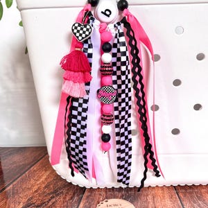 K&ouml;nnte beinhalten: Ein wei&szlig;er Tragetaschen-Anh&auml;nger mit rosa, schwarzen und wei&szlig;en Akzenten. Der Anh&auml;nger hat ein herzf&ouml;rmiges Schachbrettmuster, eine rosa Quaste und eine Perlenkette mit dem Wort "Mama". B&auml;nder und Perlen in verschiedenen Farben und Mustern sind befestigt.