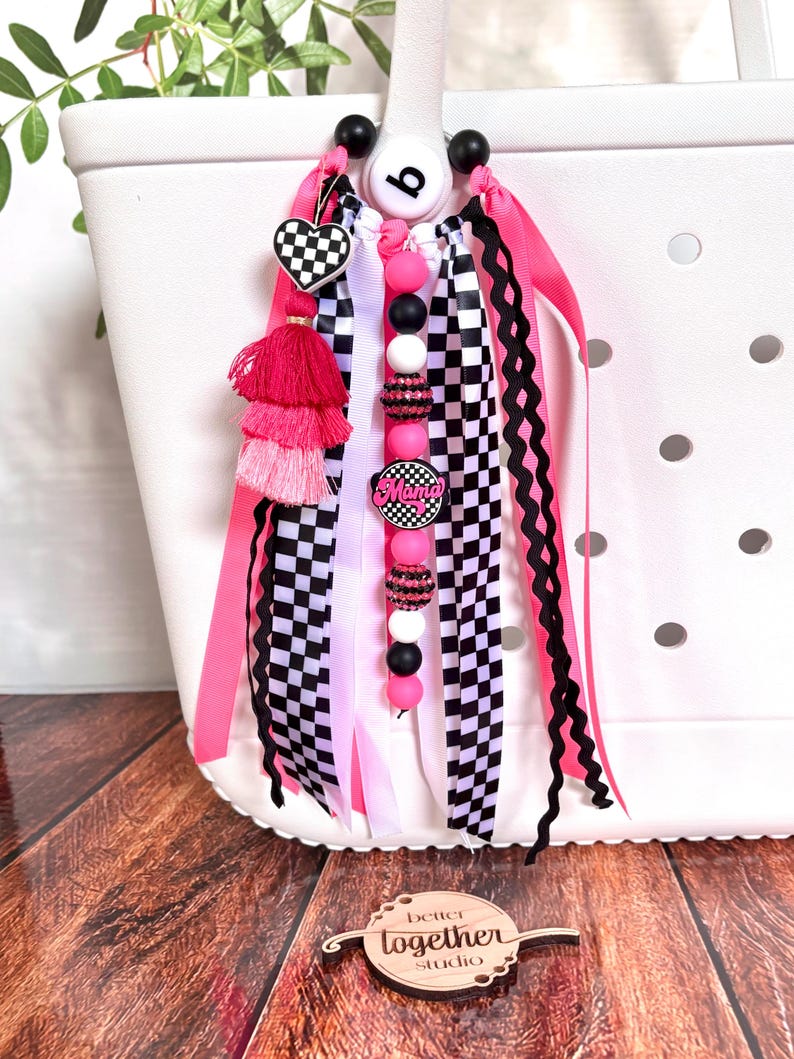 K&ouml;nnte beinhalten: Ein wei&szlig;er Tragetaschen-Anh&auml;nger mit rosa, schwarzen und wei&szlig;en Akzenten. Der Anh&auml;nger hat ein herzf&ouml;rmiges Schachbrettmuster, eine rosa Quaste und eine Perlenkette mit dem Wort "Mama". B&auml;nder und Perlen in verschiedenen Farben und Mustern sind befestigt.