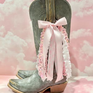May include: A grey leather cowboy boot adorned with a decorative accessory featuring a large light pink satin bow, dangling ribbons, white ruffled trim, and pink sequined lace. The boot has a pointed toe, a low heel, and intricate stitching.