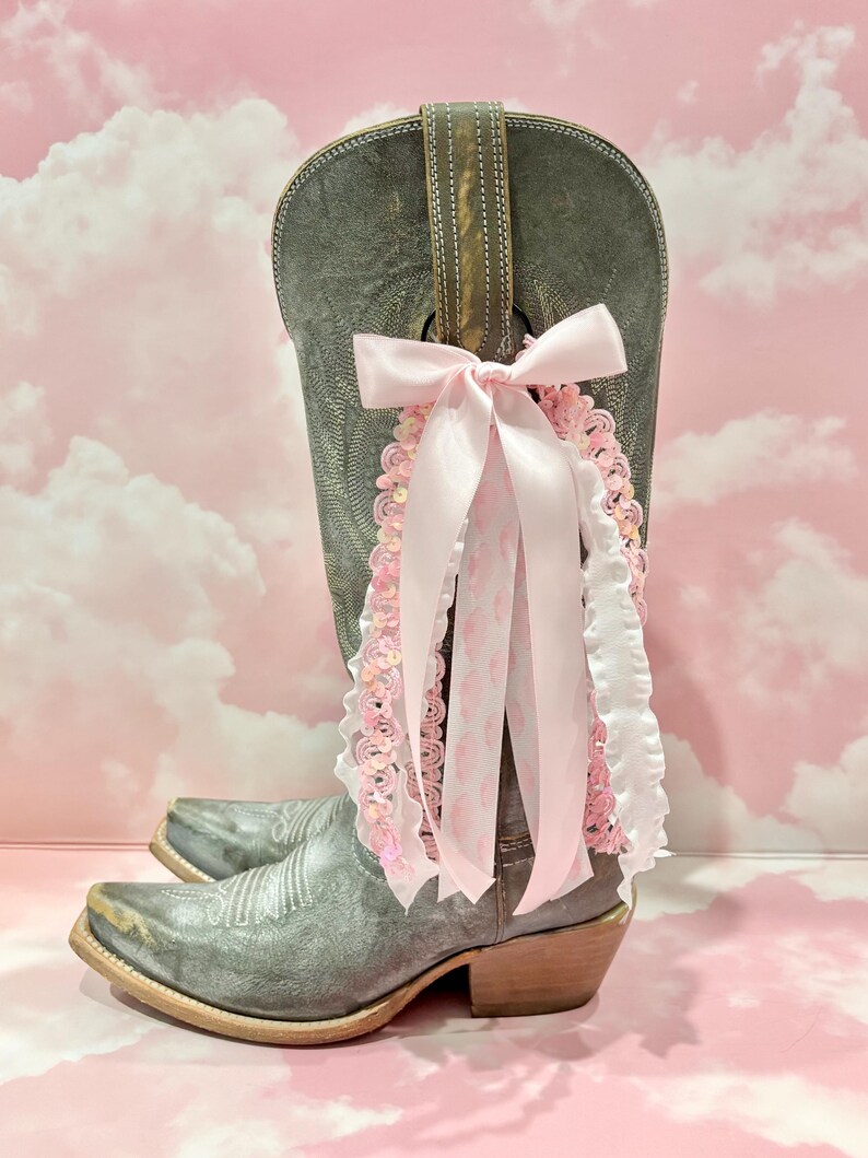 May include: A grey leather cowboy boot adorned with a decorative accessory featuring a large light pink satin bow, dangling ribbons, white ruffled trim, and pink sequined lace. The boot has a pointed toe, a low heel, and intricate stitching.
