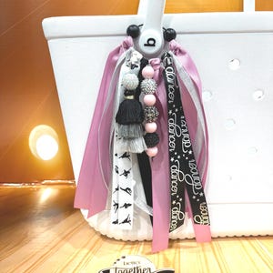 May include: White tote bag with a decorative charm. The charm includes pink and silver ribbons, black tassels, and beaded accents. A black ribbon with the word "dancer" is also attached. The bag has a white handle and a logo that says "better together studio."