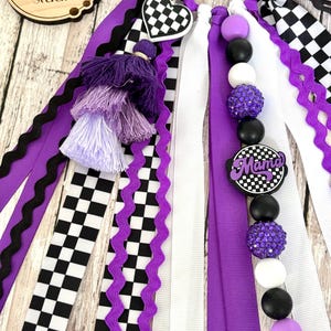 K&ouml;nnte beinhalten: Ein dekoratives Accessoire mit lila, schwarzen und wei&szlig;en B&auml;ndern, Schachbrettmustern und Perlenakzenten. Enth&auml;lt einen herzf&ouml;rmigen Anh&auml;nger und einen Anh&auml;nger mit der Aufschrift "Mama". Das Holzschild im Hintergrund tr&auml;gt die Aufschrift "better together studio".