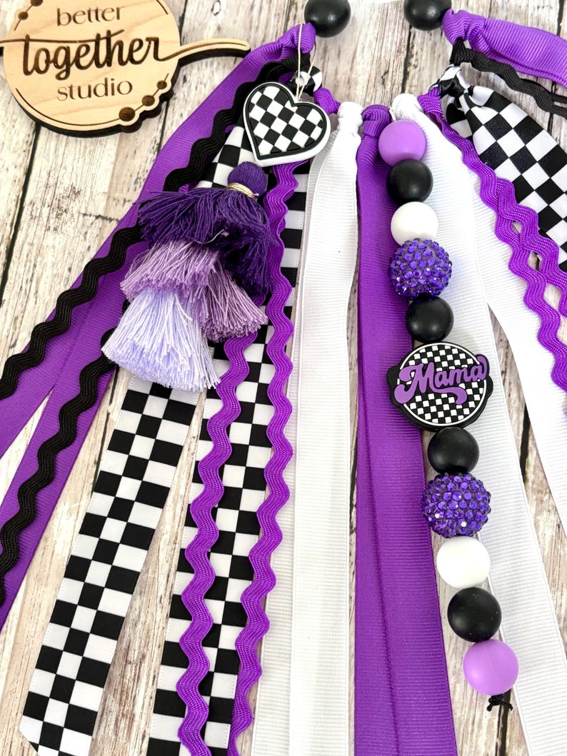 K&ouml;nnte beinhalten: Ein dekoratives Accessoire mit lila, schwarzen und wei&szlig;en B&auml;ndern, Schachbrettmustern und Perlenakzenten. Enth&auml;lt einen herzf&ouml;rmigen Anh&auml;nger und einen Anh&auml;nger mit der Aufschrift "Mama". Das Holzschild im Hintergrund tr&auml;gt die Aufschrift "better together studio".