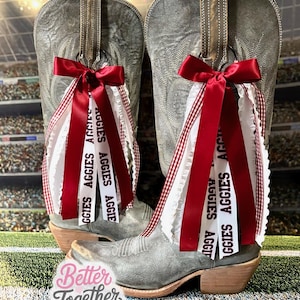 May include: A pair of gray cowboy boots with decorative red and white ribbons. The ribbons are tied with a red bow and feature the word "AGGIES". The boots have a western style with a pointed toe and a stacked heel.