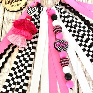 K&ouml;nnte beinhalten: Eine Sammlung farbenfroher Haaraccessoires, darunter rosa, wei&szlig;e und karierte B&auml;nder, Quasten und Perlen. Die Accessoires verf&uuml;gen &uuml;ber einen herzf&ouml;rmigen Anh&auml;nger und einen Anh&auml;nger mit der Aufschrift "Mama". Das Holzschild im Hintergrund tr&auml;gt die Aufschrift "better together studio".
