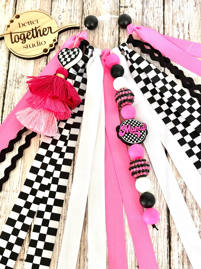 K&ouml;nnte beinhalten: Eine Sammlung farbenfroher Haaraccessoires, darunter rosa, wei&szlig;e und karierte B&auml;nder, Quasten und Perlen. Die Accessoires verf&uuml;gen &uuml;ber einen herzf&ouml;rmigen Anh&auml;nger und einen Anh&auml;nger mit der Aufschrift "Mama". Das Holzschild im Hintergrund tr&auml;gt die Aufschrift "better together studio".