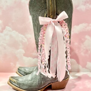 May include: A distressed grey leather cowboy boot adorned with a decorative cluster of ribbons and lace. The embellishment features a large pale pink satin bow with cascading strands of pink crochet trim, white ruffled lace, and patterned ribbon.