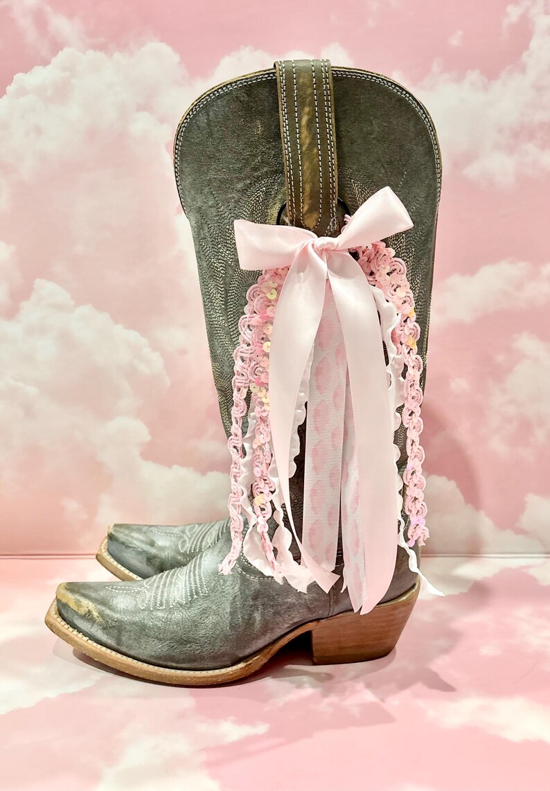 May include: A distressed grey leather cowboy boot adorned with a decorative cluster of ribbons and lace. The embellishment features a large pale pink satin bow with cascading strands of pink crochet trim, white ruffled lace, and patterned ribbon.