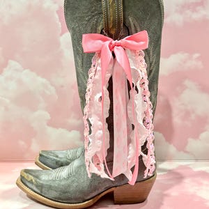 May include: A tall, distressed grey leather cowboy boot featuring a decorative pink satin bow tied to the pull strap. The bow is adorned with multiple hanging ribbons, including white ruffled trim and pink sequined lace, creating a layered effect.