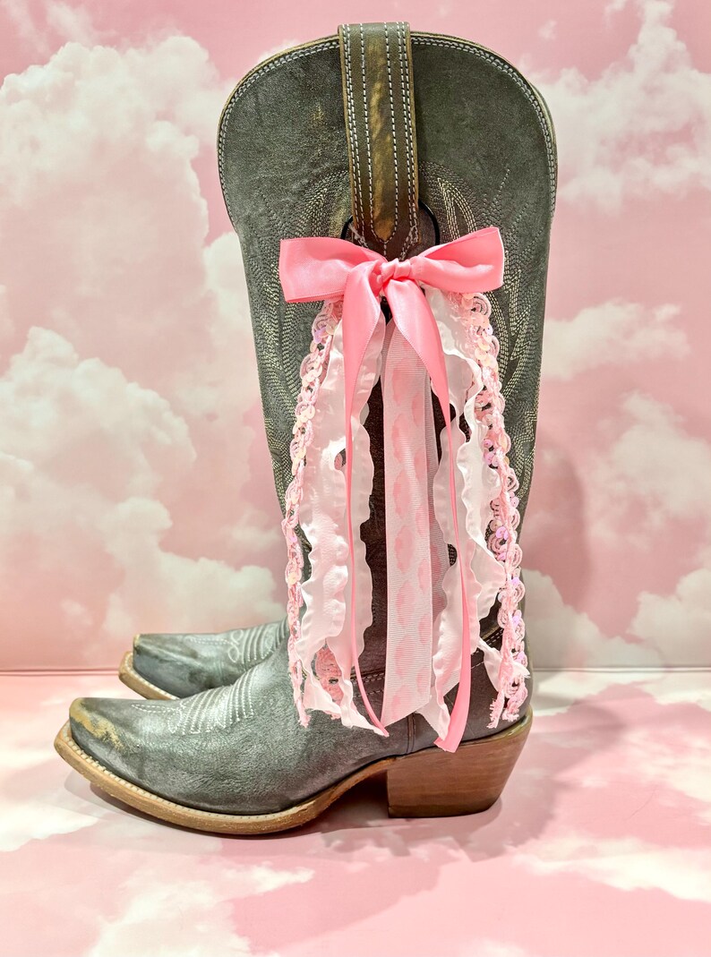 May include: A tall, distressed grey leather cowboy boot featuring a decorative pink satin bow tied to the pull strap. The bow is adorned with multiple hanging ribbons, including white ruffled trim and pink sequined lace, creating a layered effect.
