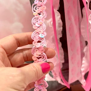 May include: A decorative boot accessory featuring a strand of light pink wavy trim adorned with iridescent sequins. The accessory includes multiple hanging ribbons in shades of bright pink and white, with some ribbons displaying a subtle pink pattern.