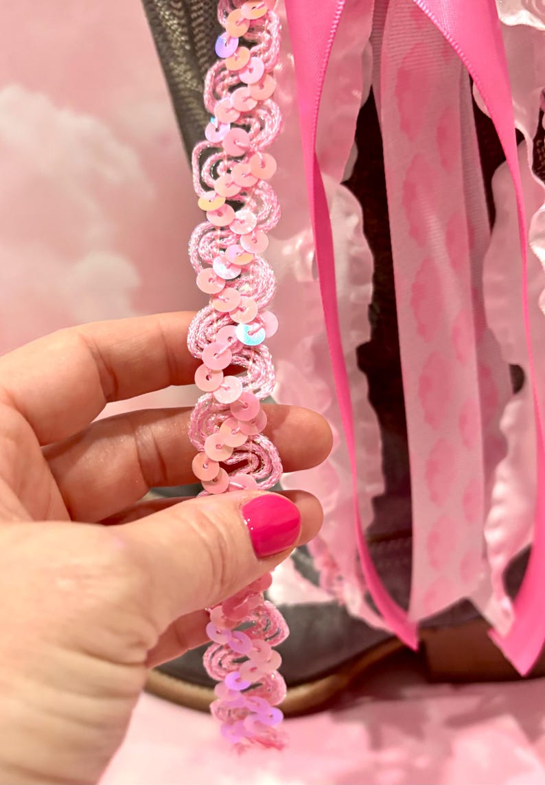 May include: A decorative boot accessory featuring a strand of light pink wavy trim adorned with iridescent sequins. The accessory includes multiple hanging ribbons in shades of bright pink and white, with some ribbons displaying a subtle pink pattern.
