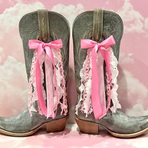May include: A pair of grey leather cowboy boots featuring a pointed toe and a stacked heel. Each boot is adorned with a decorative pink satin bow tied to the pull strap, with cascading pink and white ruffled lace ribbons hanging down the side.