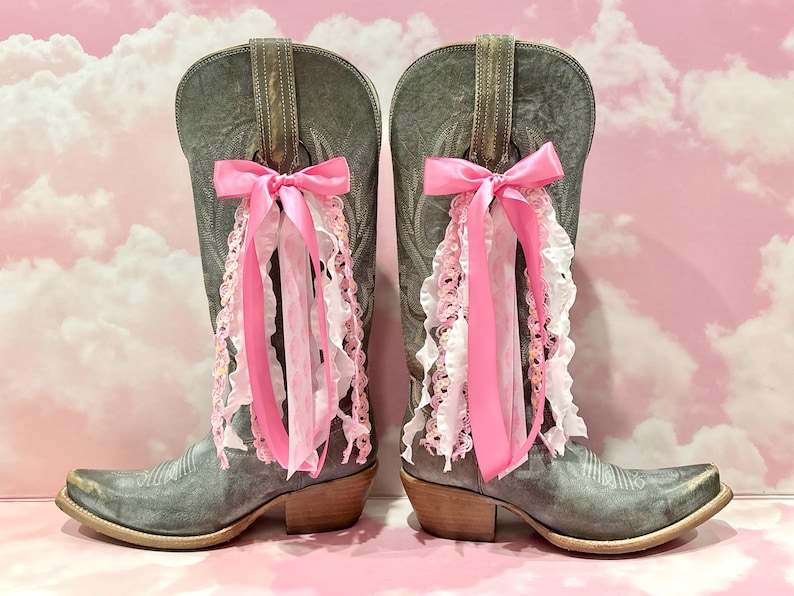 May include: A pair of grey leather cowboy boots featuring a pointed toe and a stacked heel. Each boot is adorned with a decorative pink satin bow tied to the pull strap, with cascading pink and white ruffled lace ribbons hanging down the side.