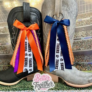 May include: Two cowboy boots, one black and one gray, are adorned with decorative ribbons. The black boot features orange and purple ribbons, while the gray boot has blue ribbons. The ribbons are printed with the word "TIGERS" and paw prints.