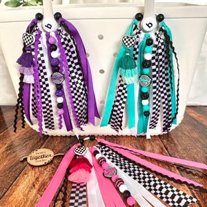 K&ouml;nnte beinhalten: Wei&szlig;e Tragetaschen mit farbenfrohen Accessoires. Die Accessoires umfassen karierte B&auml;nder, herzf&ouml;rmige Charms, Quasten und Perlenstr&auml;nge in Lila, T&uuml;rkis, Rosa und Schwarz. Die Accessoires sind an den Griffen der Taschen befestigt.