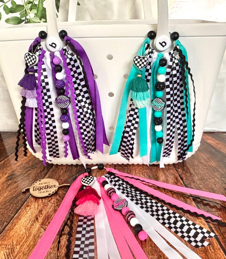 K&ouml;nnte beinhalten: Wei&szlig;e Tragetaschen mit farbenfrohen Accessoires. Die Accessoires umfassen karierte B&auml;nder, herzf&ouml;rmige Charms, Quasten und Perlenstr&auml;nge in Lila, T&uuml;rkis, Rosa und Schwarz. Die Accessoires sind an den Griffen der Taschen befestigt.