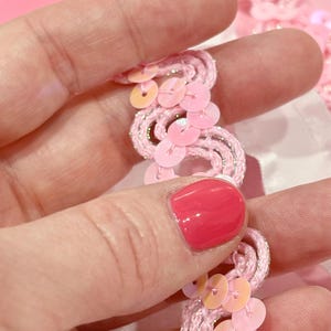 May include: A decorative trim featuring a wavy, light pink cord interwoven with thin silver metallic thread. The trim is embellished with small, round, iridescent light pink sequins that are stitched along the curves of the wavy cord structure.