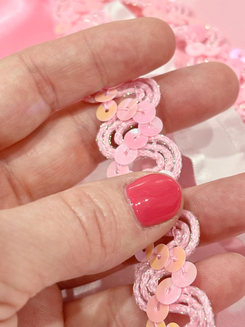 May include: A decorative trim featuring a wavy, light pink cord interwoven with thin silver metallic thread. The trim is embellished with small, round, iridescent light pink sequins that are stitched along the curves of the wavy cord structure.