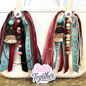 May include: Two decorative bag charms attached to a white tote bag. Each charm features a mix of maroon, black, and turquoise ribbons, lace, wooden beads, a tiered tassel, and a small animal figurine. The text Better Together Studio is visible.