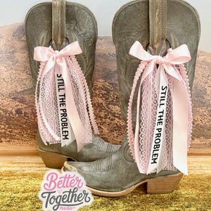 May include: A pair of gray cowboy boots adorned with pink lace and ribbons. The ribbons are tied in bows and feature the text "STILL THE PROBLEM." The boots are set against a backdrop of a landscape.