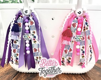 May include: Two white tote bags decorated with hanging ribbon charms in purple and pink. Each charm features a tassel, silicone beads, and ribbons printed with bingo cards, hearts, and the text Lucky Lady. The Better Together Studio logo is visible.