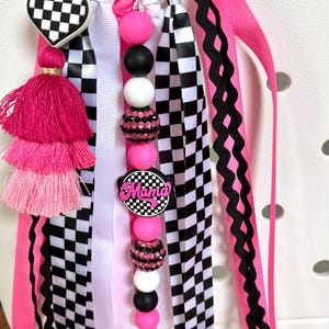 K&ouml;nnte beinhalten: Ein dekorativer Taschenanh&auml;nger mit einem herzf&ouml;rmigen Schachbrettmuster, einem rosa Quastenanh&auml;nger und einer Reihe von rosa, schwarzen und wei&szlig;en Perlen. Der Anh&auml;nger enth&auml;lt rosa, schwarze, wei&szlig;e und karierte B&auml;nder mit dem Wort "Mama" auf einem runden Anh&auml;nger.