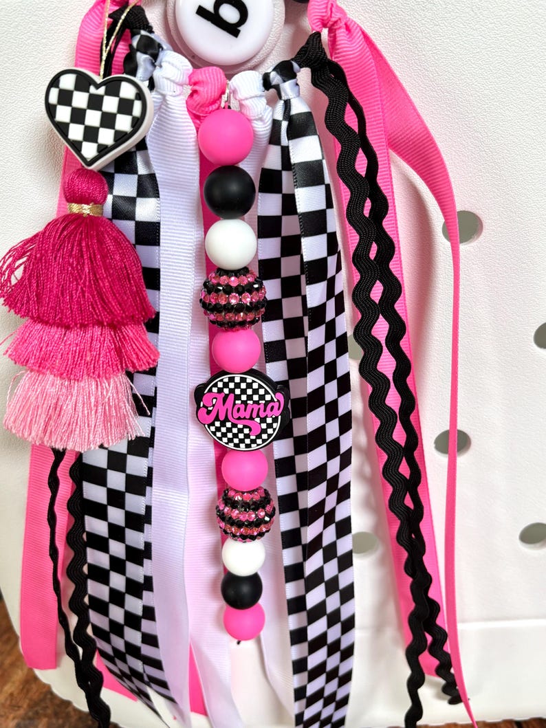 K&ouml;nnte beinhalten: Ein dekorativer Taschenanh&auml;nger mit einem herzf&ouml;rmigen Schachbrettmuster, einem rosa Quastenanh&auml;nger und einer Reihe von rosa, schwarzen und wei&szlig;en Perlen. Der Anh&auml;nger enth&auml;lt rosa, schwarze, wei&szlig;e und karierte B&auml;nder mit dem Wort "Mama" auf einem runden Anh&auml;nger.