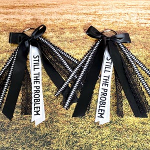 May include: Two keychains with black bows and ribbons. Each keychain has a white ribbon printed with "STILL THE PROBLEM" in black. The ribbons are accented with black lace and black and white striped ribbons. The background is a field of dry grass.