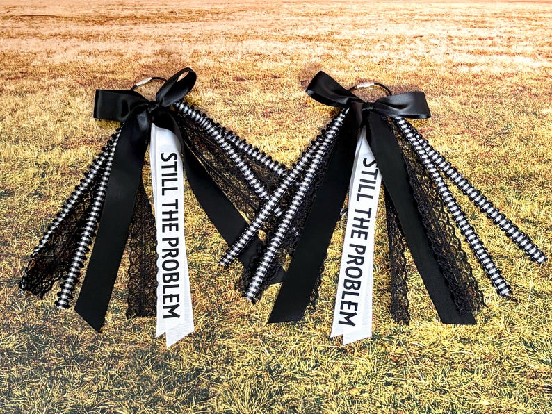 May include: Two keychains with black bows and ribbons. Each keychain has a white ribbon printed with "STILL THE PROBLEM" in black. The ribbons are accented with black lace and black and white striped ribbons. The background is a field of dry grass.