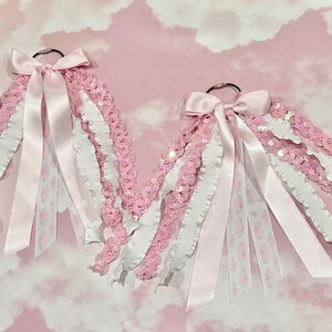 May include: Two decorative hanging ornaments featuring silver metal rings adorned with cascading pink and white ribbons, lace trim, and sequined fabric strips. Each ornament is finished with a satin ribbon bow at the top, creating a layered, textured appearance with various fabric widths and patterns.