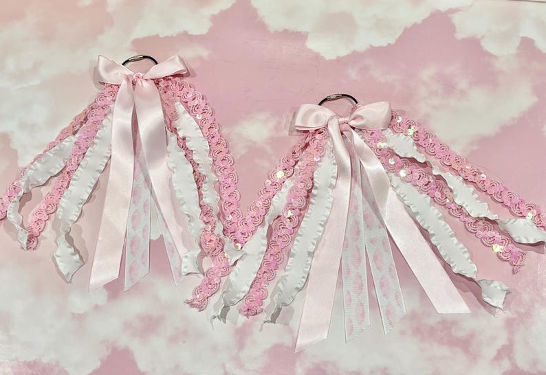 May include: Two decorative hanging ornaments featuring silver metal rings adorned with cascading pink and white ribbons, lace trim, and sequined fabric strips. Each ornament is finished with a satin ribbon bow at the top, creating a layered, textured appearance with various fabric widths and patterns.
