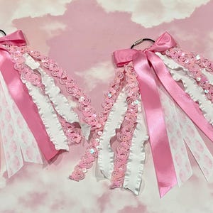 May include: Two identical cheerleading hair bows attached to silver metal rings. Each bow features a combination of pink satin ribbons, white ruffled ribbons, and pink sequined trim, all layered together to create a full, textured appearance.