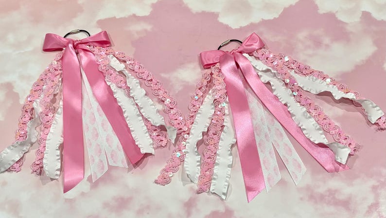 May include: Two identical cheerleading hair bows attached to silver metal rings. Each bow features a combination of pink satin ribbons, white ruffled ribbons, and pink sequined trim, all layered together to create a full, textured appearance.