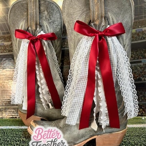 May include: A pair of gray cowboy boots adorned with red satin bows and white lace and gingham accents. The boots have a distressed look and are decorated with a decorative boot accessory. The text "Better Together Studio" is visible.